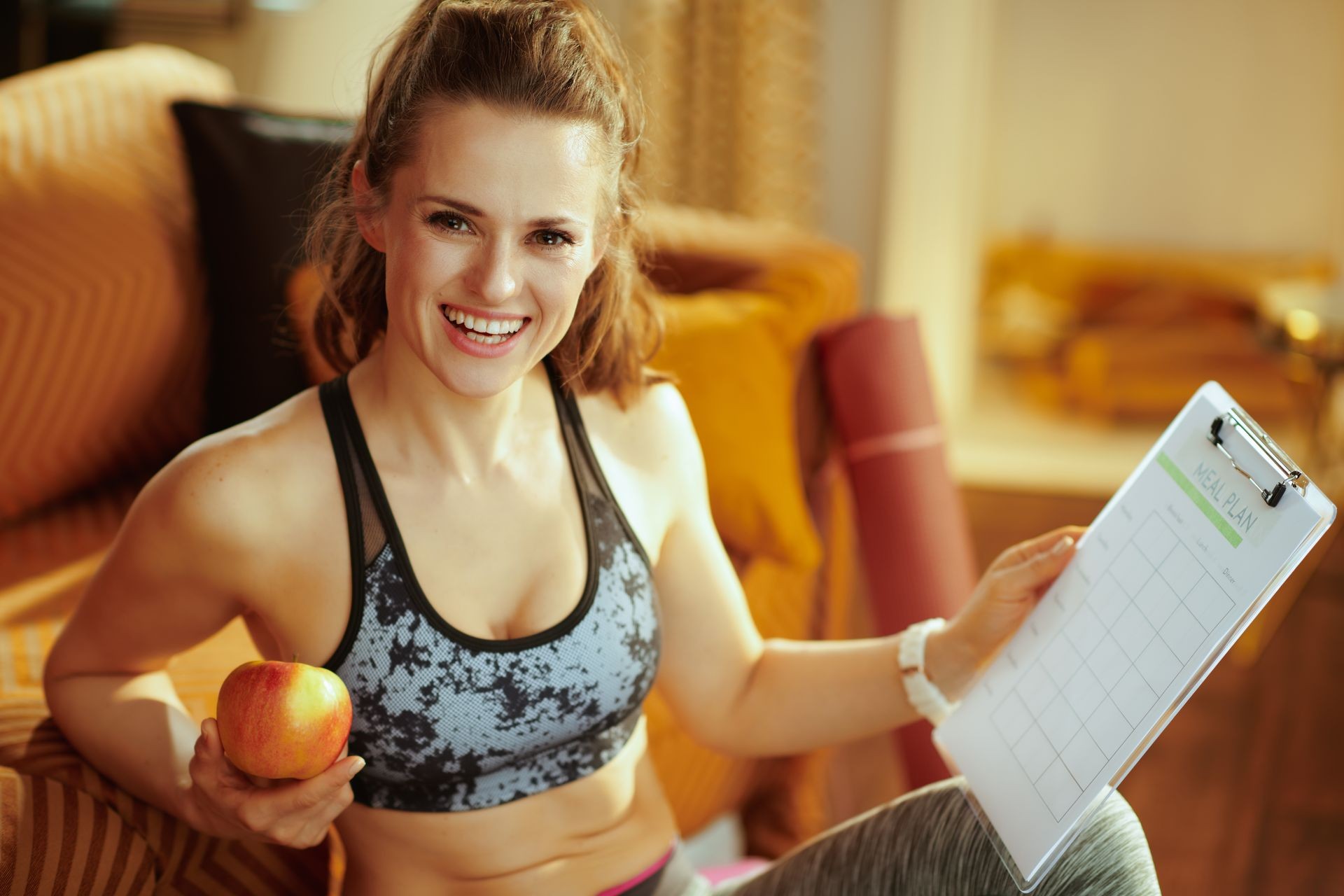 Portrait of happy young sports woman in sport clothes holding apple and clipboard with meal plan at modern home.